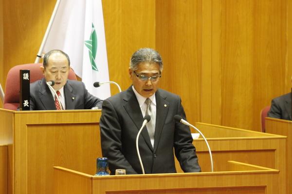 議会の壇上にて教育長が手元の資料を読みながら教育方針を話している様子の写真